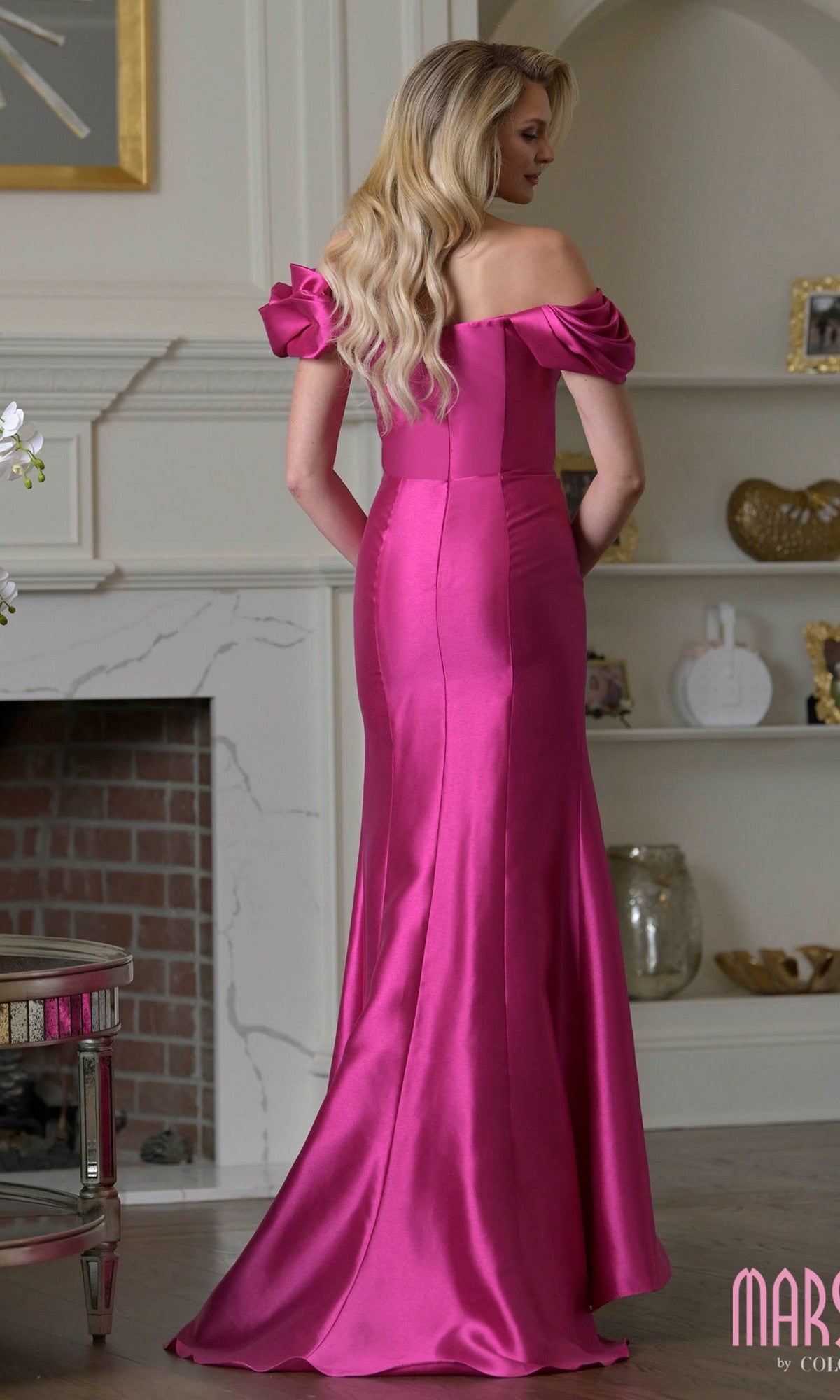 A woman with long blonde hair wears the Marsoni MV1407, a bright pink ruffled off-shoulder satin mermaid gown, standing in a room with a fireplace and shelves.